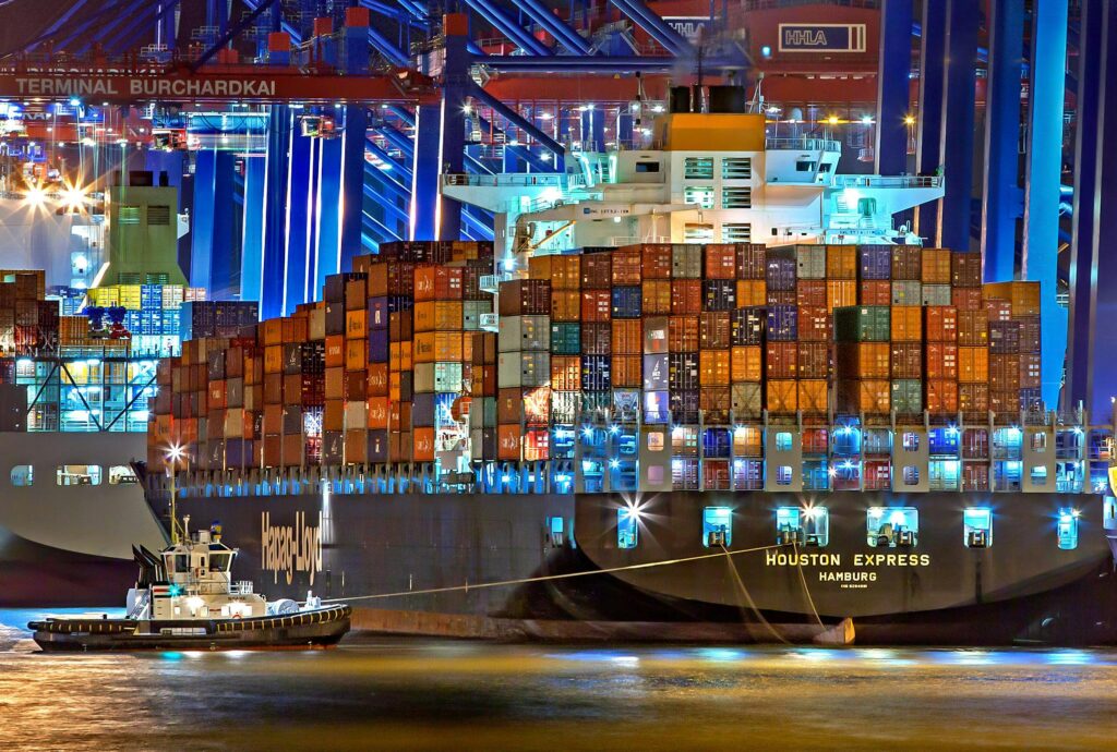 A brightly lit cargo ship at Hamburg harbor with stacked containers and a tugboat.